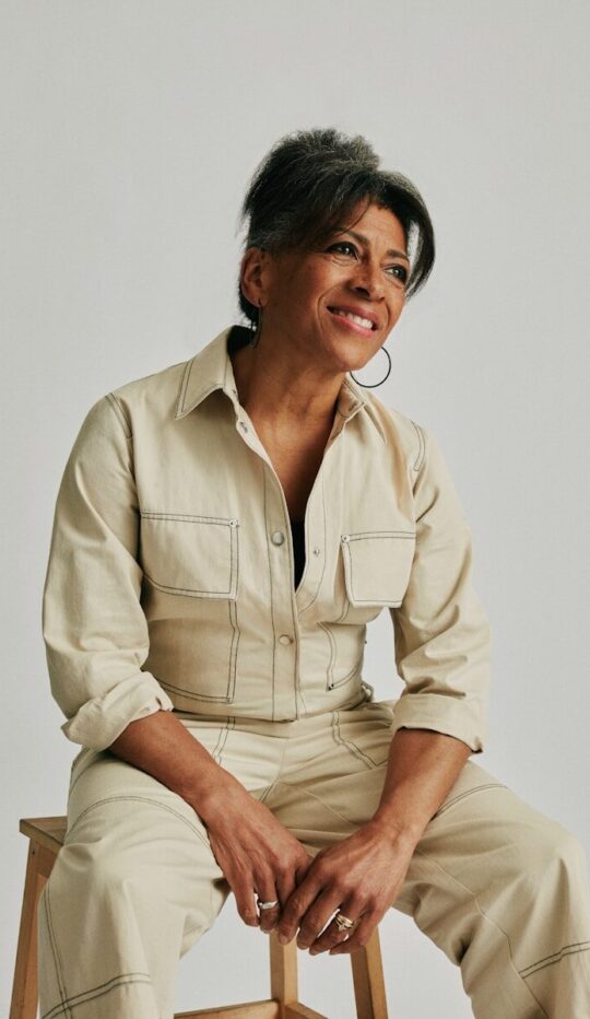 Woman in beige jumpsuit smiling, sitting on a stool.